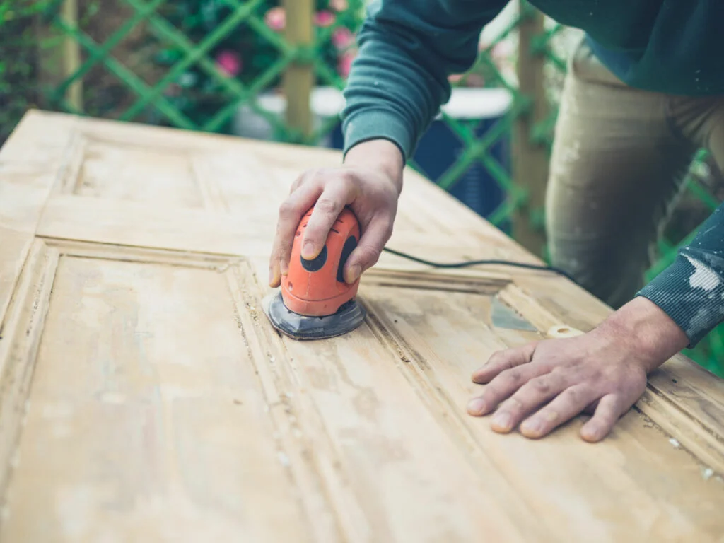 A young man is sanding an old door with an electric sander